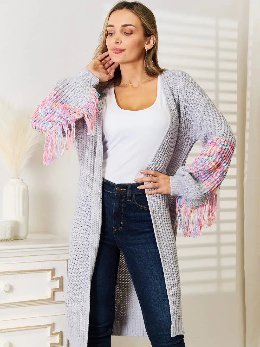 Woman wearing a long, light gray cardigan with colorful fringe sleeves in a room.