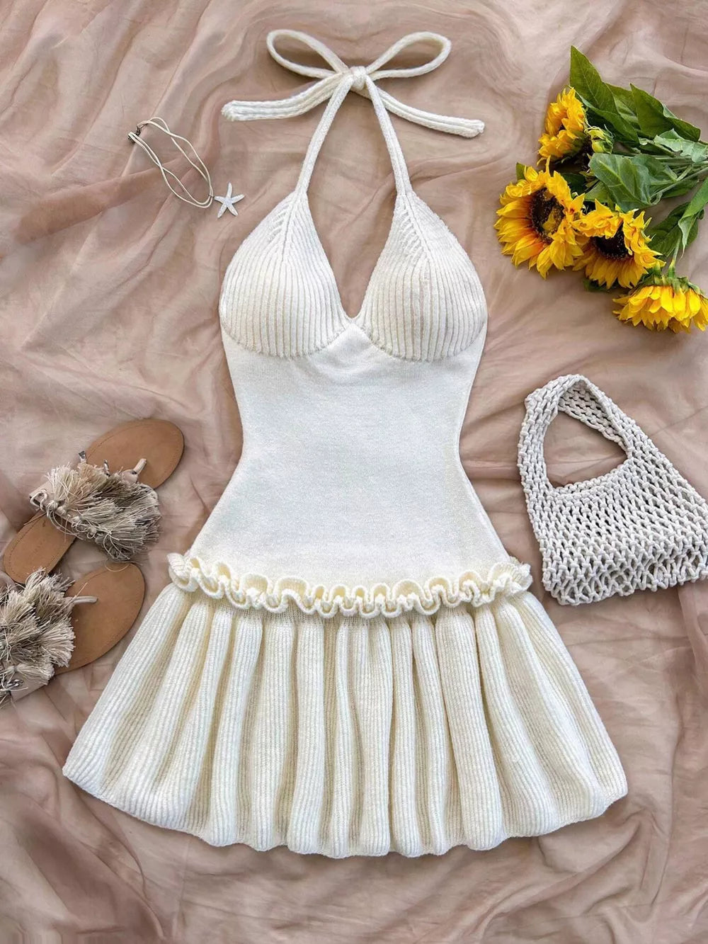 White ribbed swimsuit with ruffle hem on a beige surface with flowers and sandals.