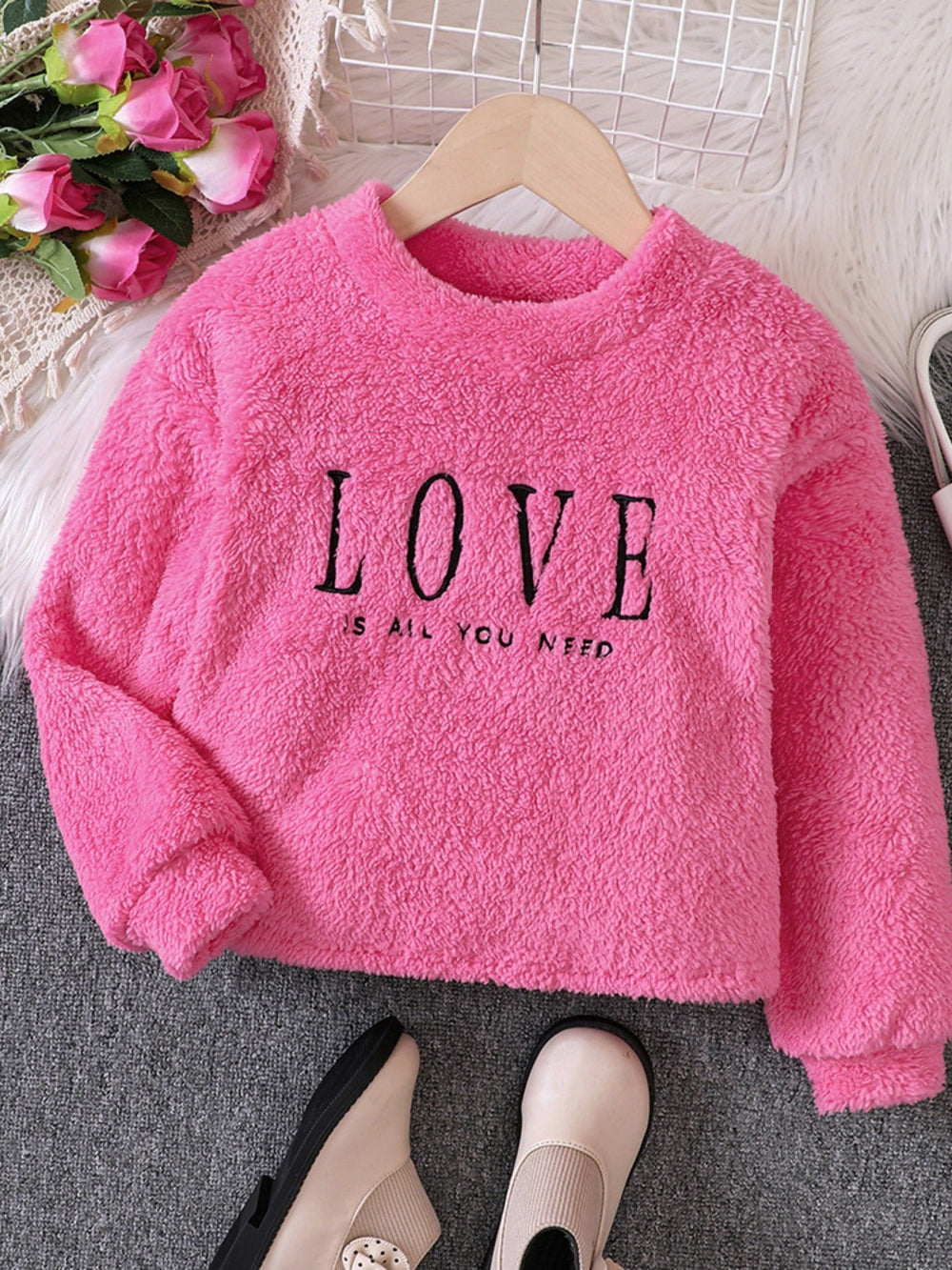 Pink fleece sweatshirt with 'LOVE IS ALL YOU NEED' text on a hanger with flowers and shoes in the background.
