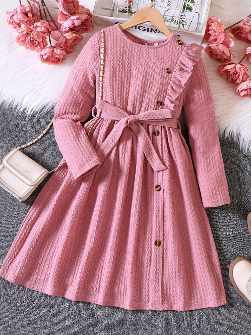 Pink dress with buttons and ruffles on a hanger with decorative flowers and a handbag in the background.