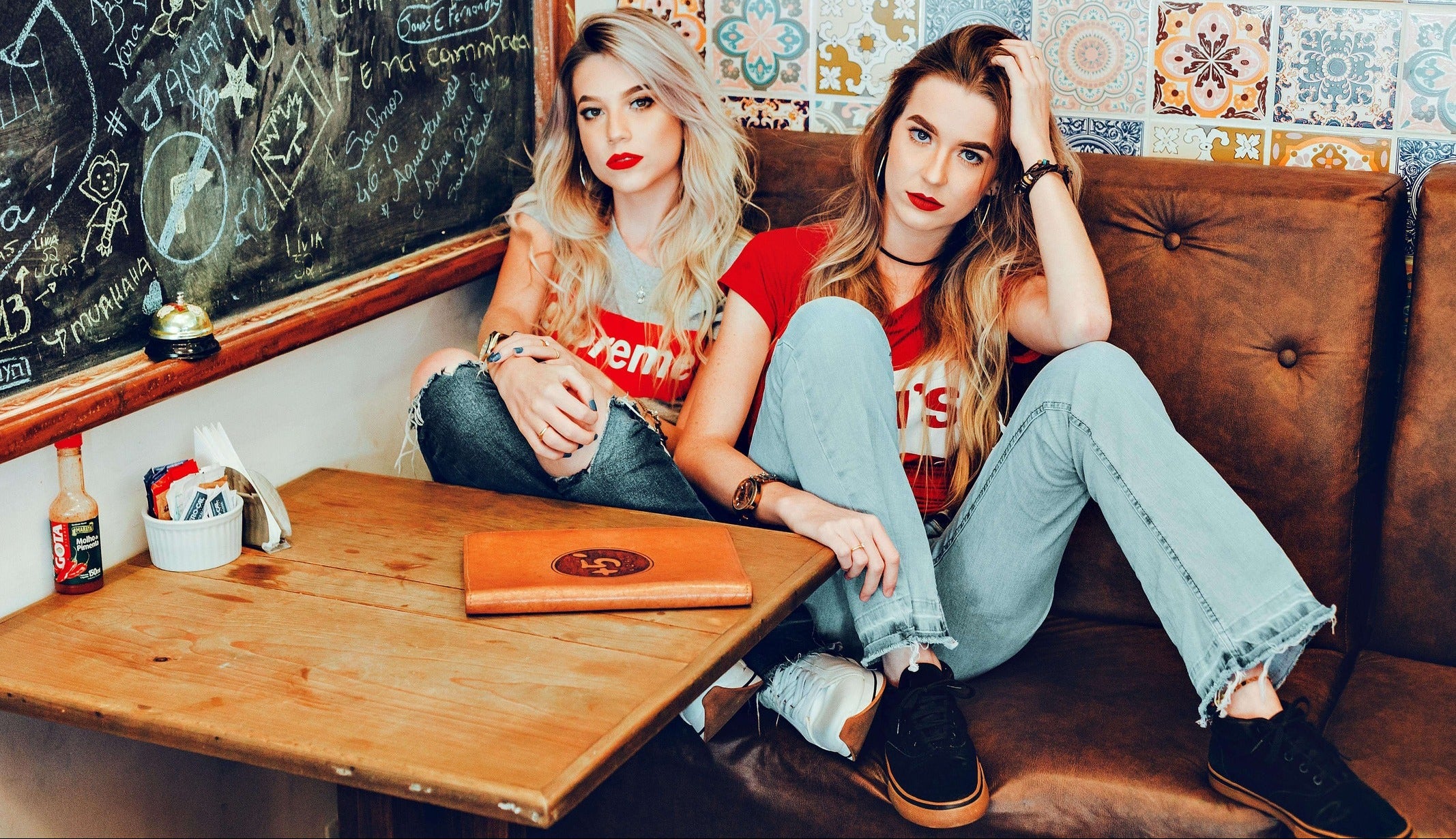 Two women sitting on a couch in a casual setting with a chalkboard wall and colorful tiles.